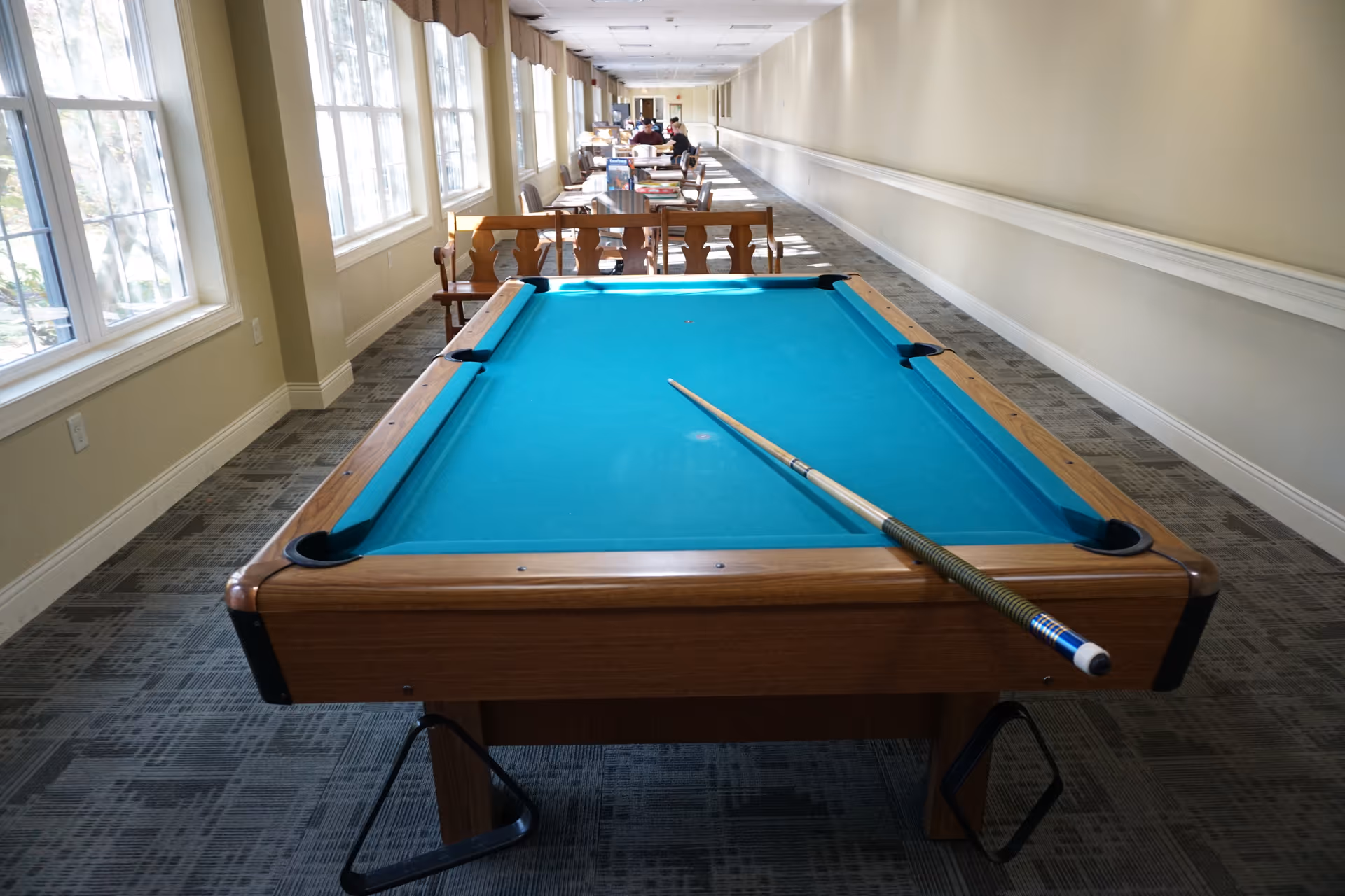 A pool table in a long, sunlit common corridor with windows on the left and tables and chairs in the background.
