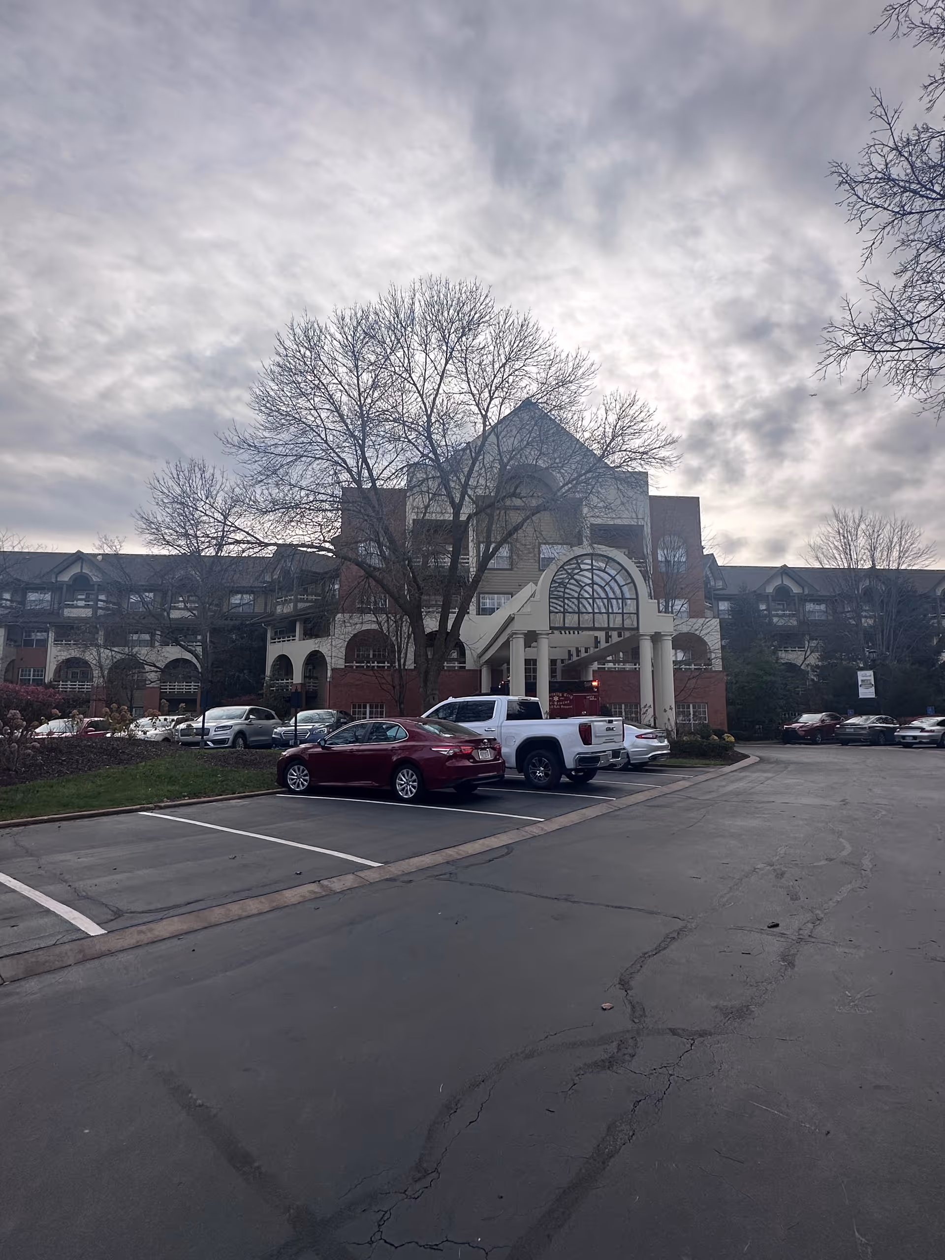 Exterior view of The Gatesworth senior living facility showing a multi-story building with a covered entrance supported by columns. Several cars are parked in the parking lot in front of the building, and leafless trees are visible around the area under a cloudy sky.