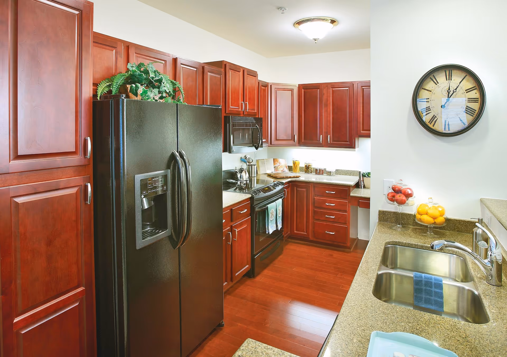 A modern kitchen with dark wood cabinets, a black refrigerator with water dispenser, a black stove with oven, a microwave, and a double sink with a blue towel. There is a round wall clock above the counter and bowls of fruit on the countertop.