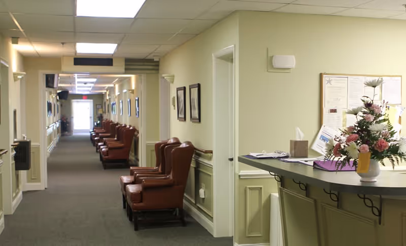 A long hallway in a senior living facility with multiple brown leather armchairs lined up against the walls. On the right side, there is a reception desk with a vase of flowers and some papers. The walls are painted light yellow, and there are framed pictures hanging along the hallway. The ceiling has fluorescent lights.