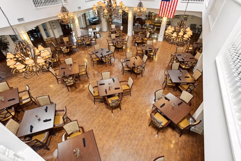 Spacious dining room with multiple square wooden tables, each surrounded by four chairs with cushioned seats and backs. The tables are set with black napkins and small floral centerpieces. Large ornate chandeliers hang from the ceiling, illuminating the room. An American flag is displayed on one wall, and the floor is made of polished wood.