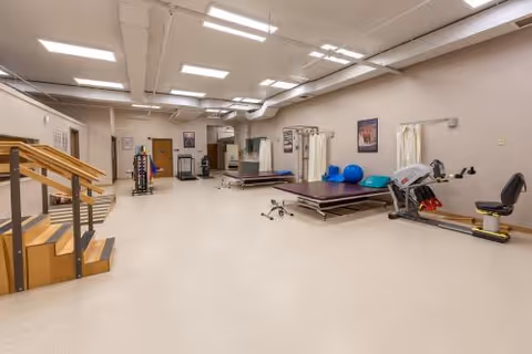 A spacious physical therapy or rehabilitation room with exercise equipment including parallel bars, therapy tables, exercise balls, and a stationary recumbent bike. The room has bright overhead lighting, beige walls, and a clean, open floor space.