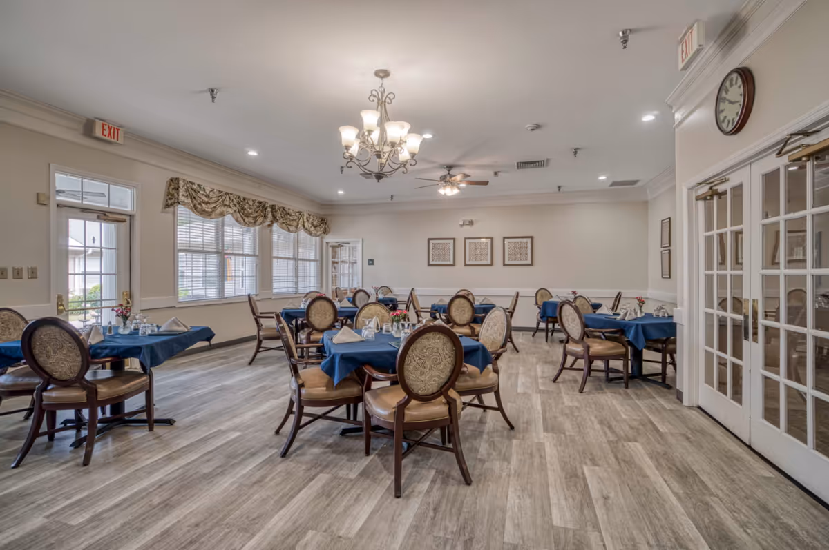 Spacious dining room with round tables set with blue tablecloths and upholstered chairs beneath chandeliers.
