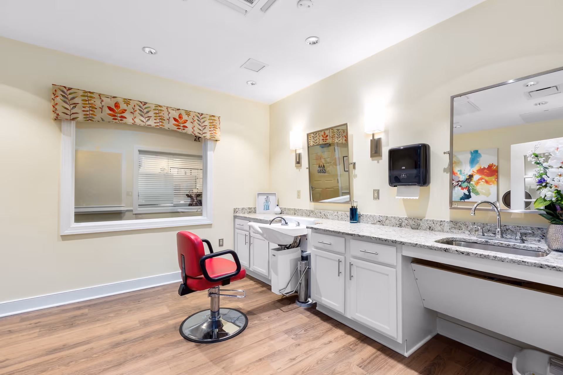 Interior view of a hair salon area in a senior living facility featuring a red salon chair in front of a white countertop with a built-in hair washing sink, a large mirror, and a paper towel dispenser mounted on the wall. The room has light yellow walls, wooden flooring, and a window with a decorative valance.