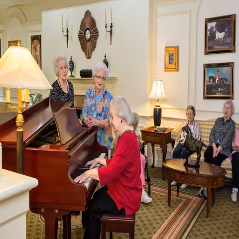 A group of elderly women in a cozy living room setting, with one woman playing a grand piano while two others stand nearby singing or listening. Three more women are seated on a couch and chair in the background, surrounded by traditional decor including lamps, framed paintings of horses, and a small table with a horse statue.