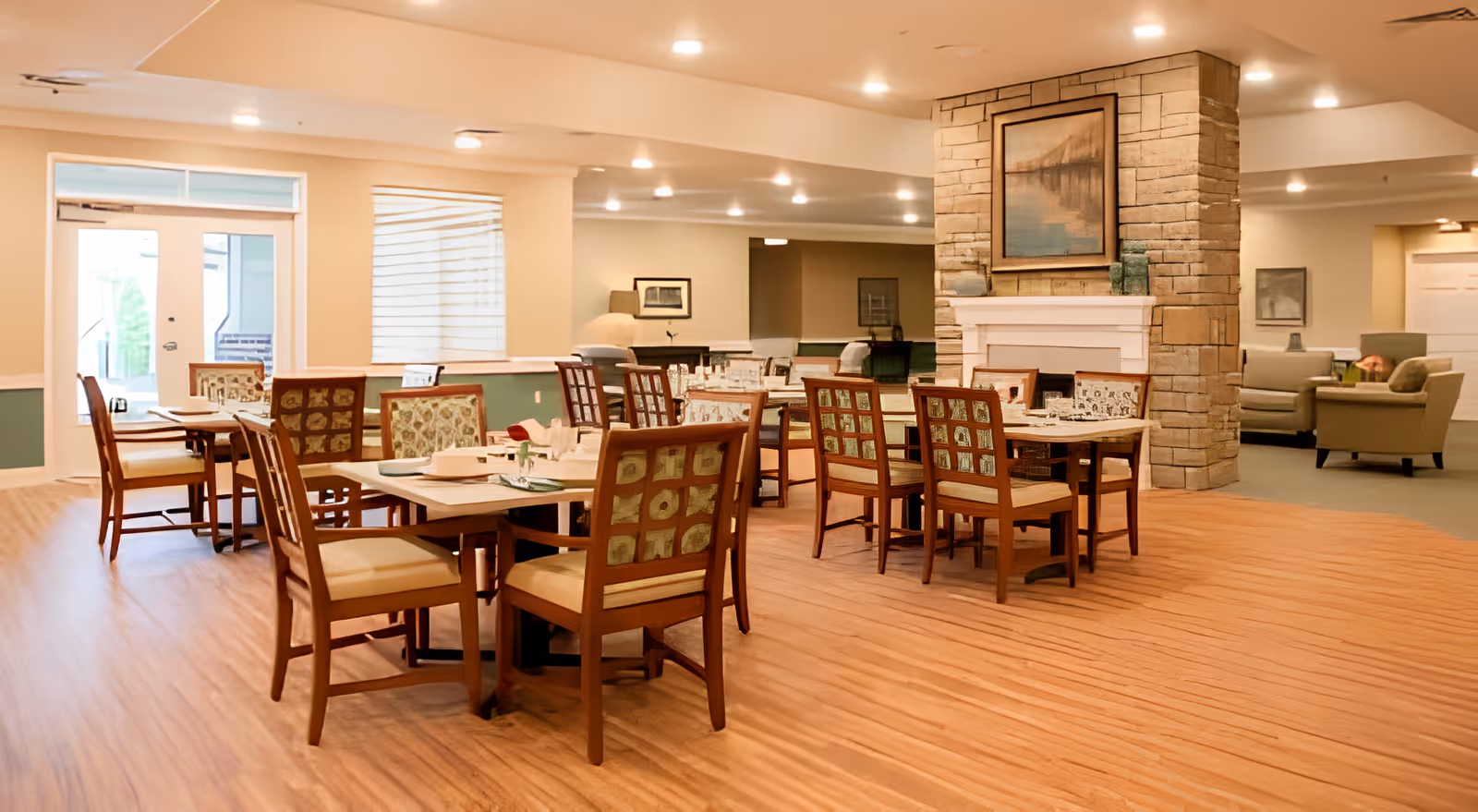 A spacious dining area in The Ganzhorn Suites featuring multiple wooden tables and chairs with patterned upholstery. The room has a warm wooden floor, a stone fireplace with a framed painting above it, and soft lighting from recessed ceiling lights. There is a seating area with armchairs in the background and large windows letting in natural light.