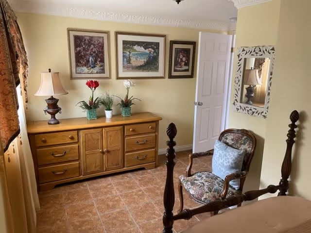 Cozy bedroom with a wooden dresser topped with a lamp and potted plants, framed art on pale yellow walls, and a carved wooden chair beside a four-poster bed.