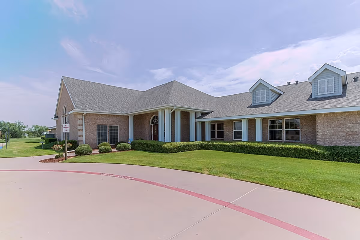 Exterior view of a single-story brick building with a gray shingled roof, white columns at the entrance, and neatly trimmed bushes and green lawn surrounding it under a partly cloudy sky.