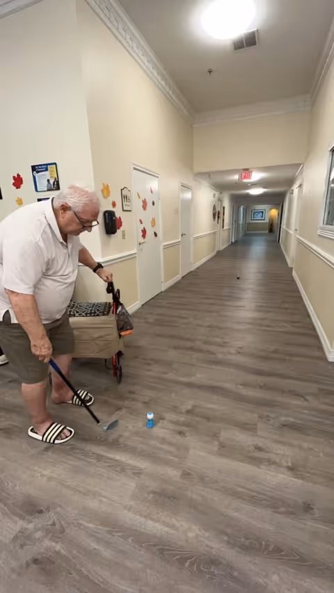 An elderly man playing indoor mini golf in a long hallway of an assisted living facility. The hallway has light-colored walls decorated with autumn leaf cutouts and several closed doors. The floor is wood-patterned, and the man is using a putter to hit a small blue ball.