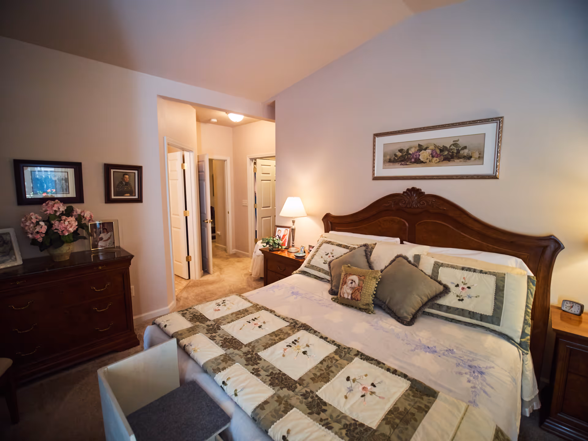 A cozy, traditionally furnished bedroom with a wooden headboard bed, floral quilt, dresser and bedside table with lamp.