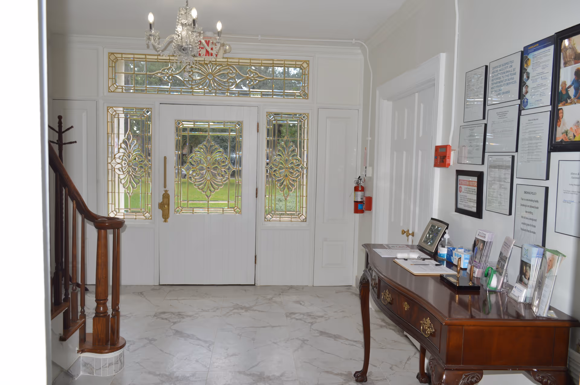 Entrance area of an assisted living facility with a decorative glass front door and sidelights. There is a wooden staircase railing on the left and a wooden console table on the right with brochures, hand sanitizer, and framed pictures. Several framed notices and documents are displayed on the wall above the table. The floor is made of light-colored marble tiles.