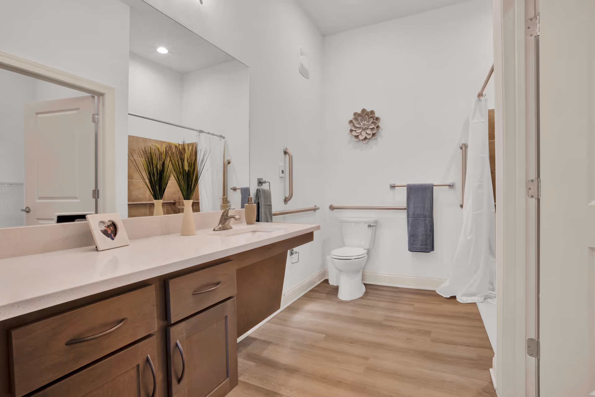 A clean and modern bathroom with a large mirror above a long countertop with a sink. There are wooden cabinets below the countertop. On the countertop, there is a small framed photo and a vase with tall green plants. The bathroom has a toilet with grab bars on the walls and a towel hanging on a bar. A shower with a white curtain is visible in the background. The floor is wood-style, and the walls are white with a decorative flower wall hanging above the toilet.