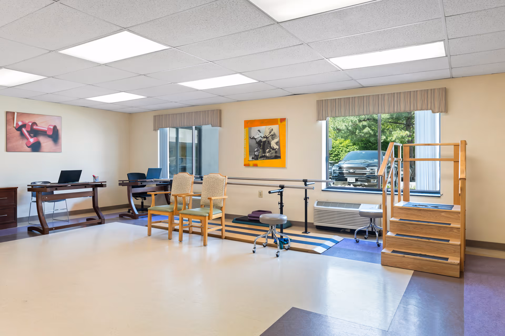 A rehabilitation therapy room with wooden stairs and handrails for physical therapy, two chairs, two desks with laptops, and large windows letting in natural light. The walls have framed pictures, one showing red dumbbells and another showing an exercise scene.