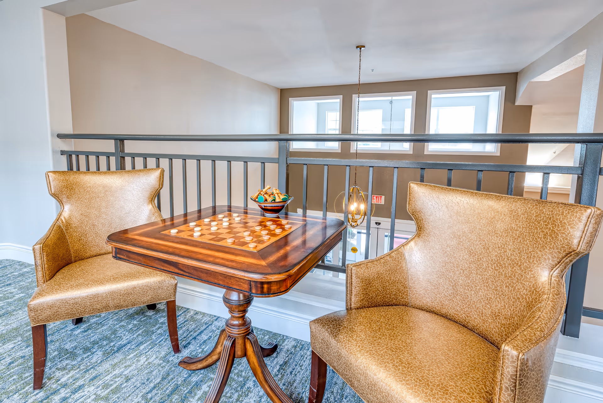 A cozy seating area with two brown leather armchairs and a wooden table with a built-in checkers board. The table has a bowl with snacks on it. The area overlooks a railing with windows and a hanging light fixture in the background.