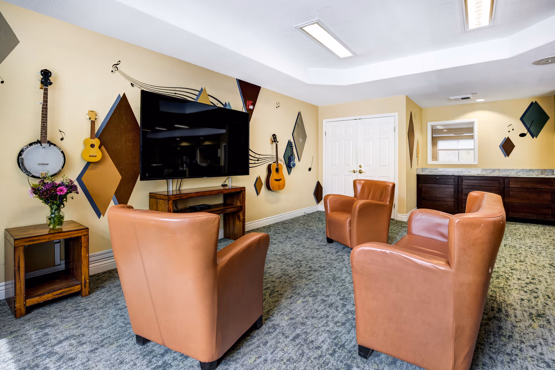 Cozy communal lounge with four leather armchairs facing a wall-mounted TV and musical instruments mounted on a decorated wall.