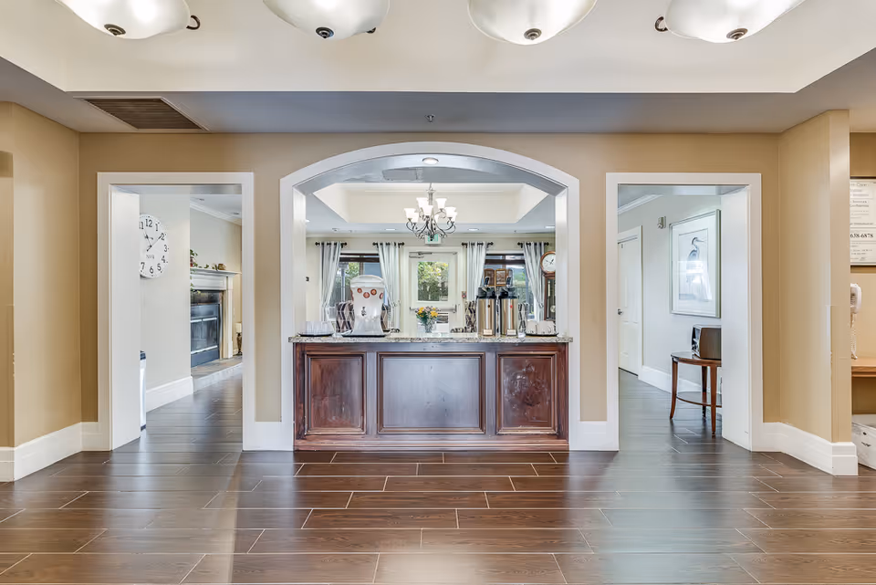 Interior common area with a wooden service counter beneath an archway, coffee urns on the counter and a view into a seating area with a chandelier.