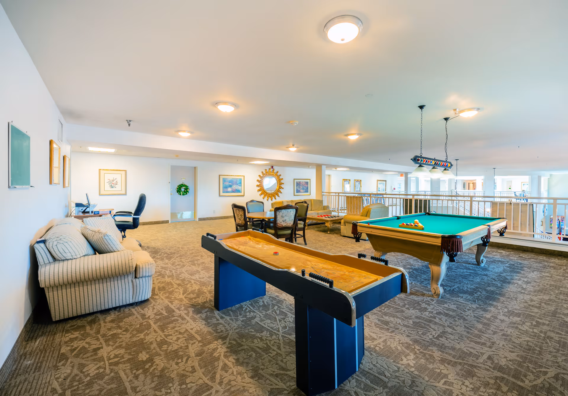 Spacious indoor recreation room with a pool table, shuffleboard, seating area, and tables in a senior living facility.