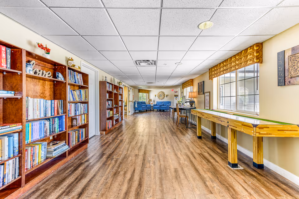 A long bright common room with bookshelves on the left, a shuffleboard table on the right, and seating at the far end.