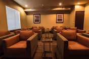 Cozy communal lounge with rows of cushioned armchairs, small side tables, framed artwork, and warm recessed lighting.