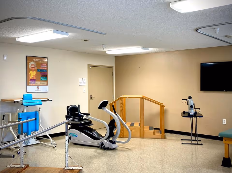 A rehabilitation room with exercise equipment including a recumbent exercise bike, parallel bars for walking practice, a small wooden staircase with handrails, and a vision testing device on a stand. There is a TV mounted on the wall and a poster about types of dementia on another wall.