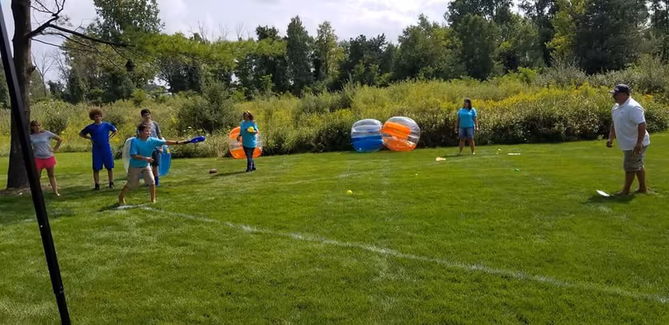 A group of people playing a lawn game on a grassy field with trees and bushes in the background. Some individuals are holding paddles and balls, and there are inflatable bumper balls on the grass.