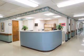 Curved blue reception desk in a well-lit interior corridor of a senior living facility with filing shelves, potted plants, and fluorescent ceiling lights.