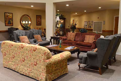 A cozy living room area in a senior living facility with multiple upholstered sofas and armchairs arranged around a wooden coffee table. The room is warmly lit with wall art, a mirror, and decorative plants adding to the inviting atmosphere.