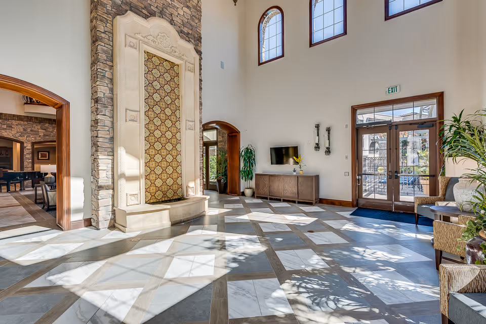 Bright and spacious senior living facility common area with high ceilings, large windows, and a decorative stone water feature. The room has patterned tile flooring, a wooden cabinet with a TV mounted above it, potted plants, and seating areas near the entrance doors.