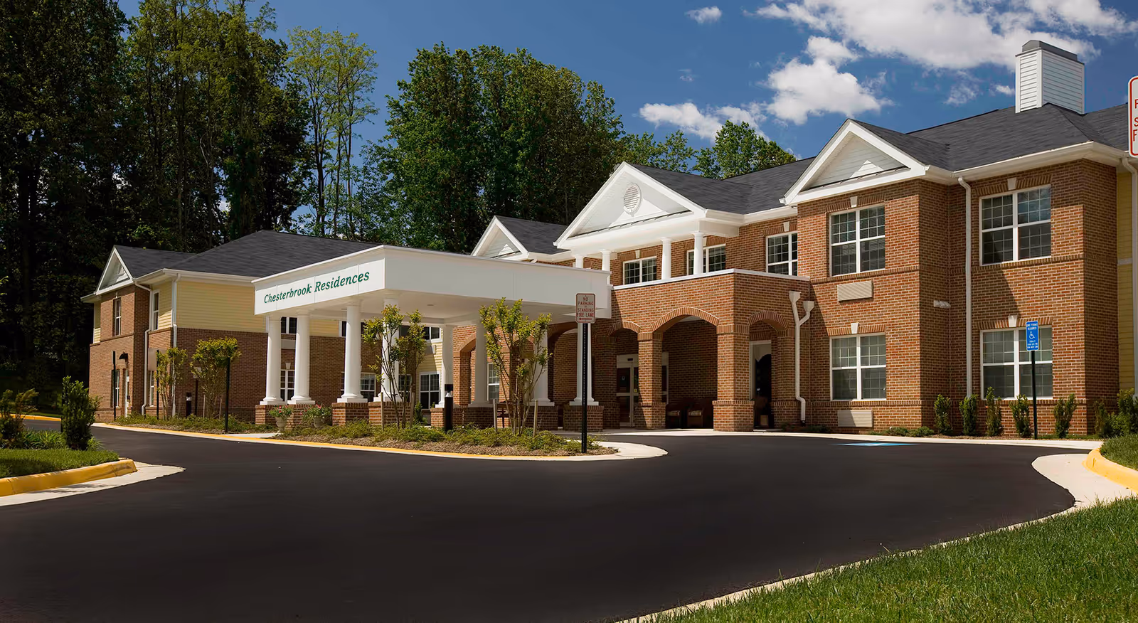 Exterior view of Chesterbrook Residences, a two-story brick building with white columns and a covered entrance. The building is surrounded by trees and landscaping under a partly cloudy sky.