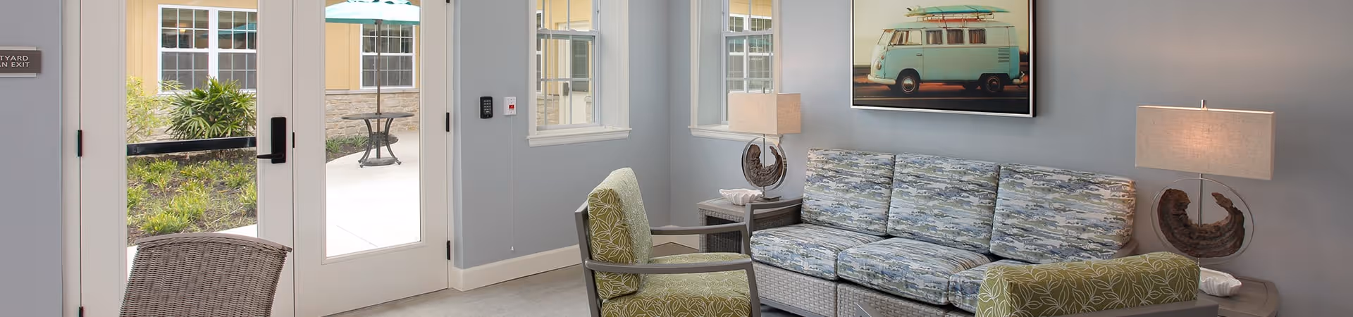 A bright and cozy living room area with a patterned sofa, two green armchairs, two side tables each with a lamp, and a framed picture of a vintage van on the wall. Large windows and glass doors provide a view of an outdoor patio with tables and greenery.