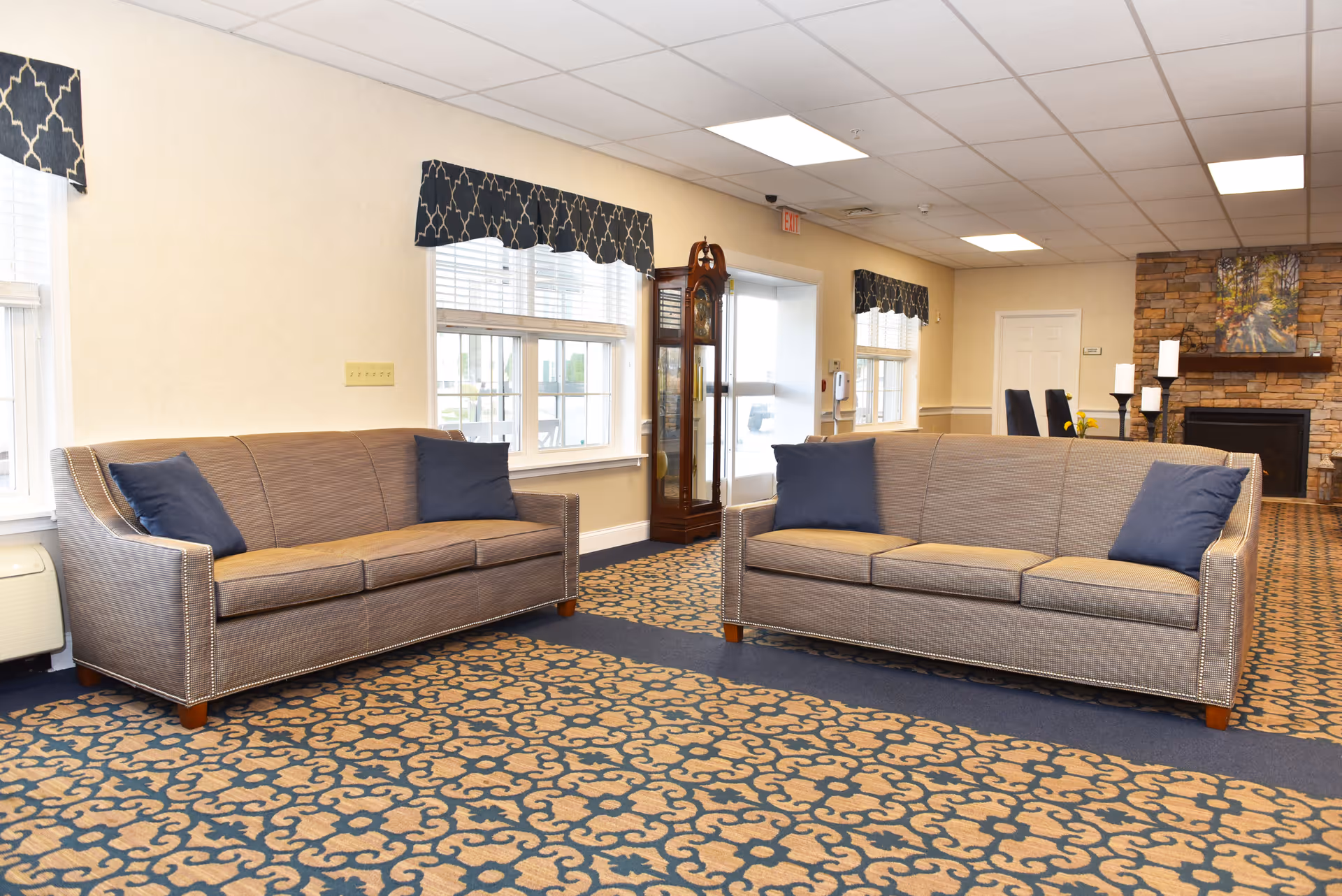 A senior living facility common area with two beige sofas each with two blue throw pillows, patterned carpet, large windows with blue valances, a grandfather clock, and a stone fireplace with candles and a painting above it.