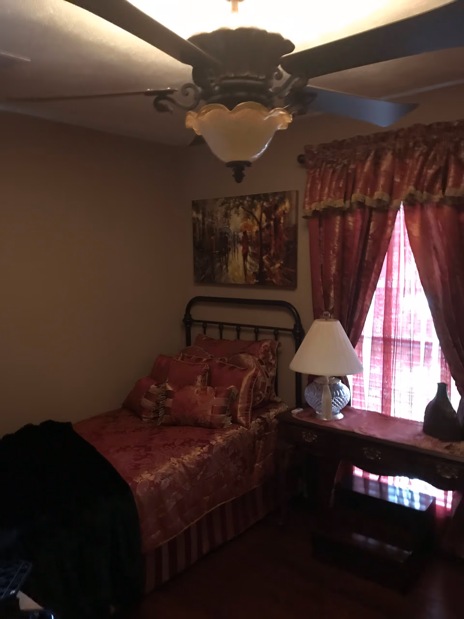 A bedroom with a single bed covered in red and gold patterned bedding and multiple pillows. There is a wooden nightstand next to the bed with a white lamp and decorative items. Red curtains with gold trim cover a window, and a painting depicting a street scene hangs on the wall above the bed. A ceiling fan with a light fixture is visible on the ceiling.