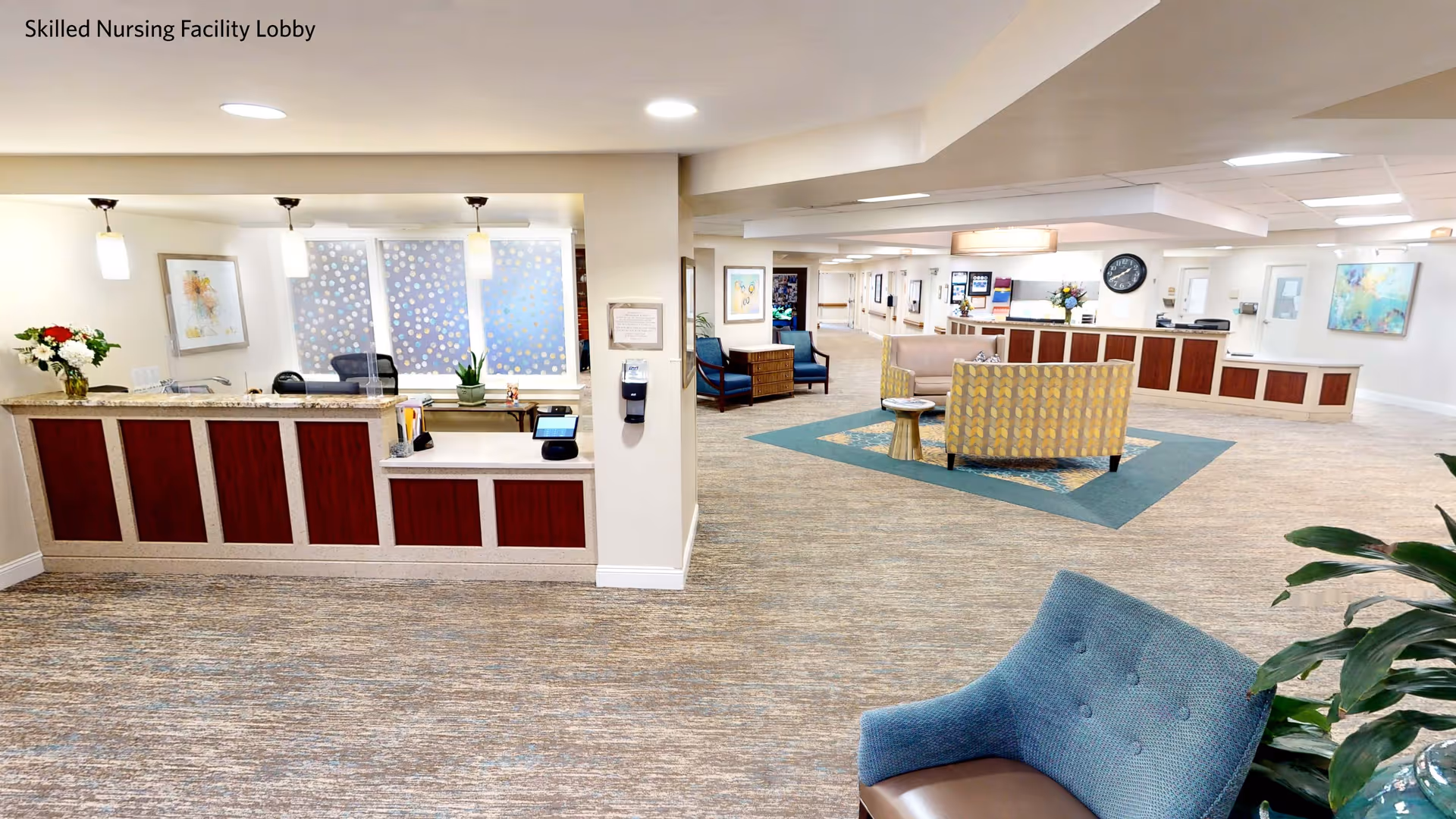 A spacious skilled nursing facility lobby with a reception desk on the left side, decorated with flowers and pendant lights. The lobby has comfortable seating areas with chairs and sofas arranged on a patterned rug. The walls are adorned with framed artwork, and there is a clock above the reception area. The space is well-lit with ceiling lights and has a clean, welcoming atmosphere.