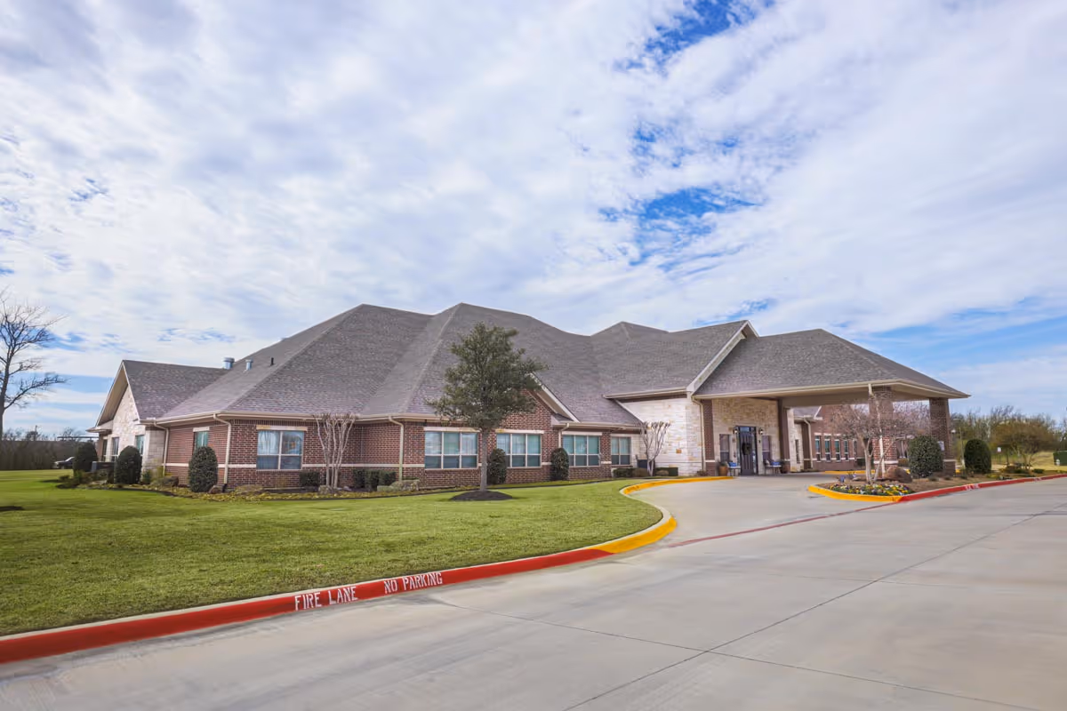 Exterior view of a single-story assisted living facility building with a brick and stone facade, surrounded by a well-maintained lawn and small trees under a partly cloudy sky. The driveway curves toward the main entrance with a covered drop-off area.