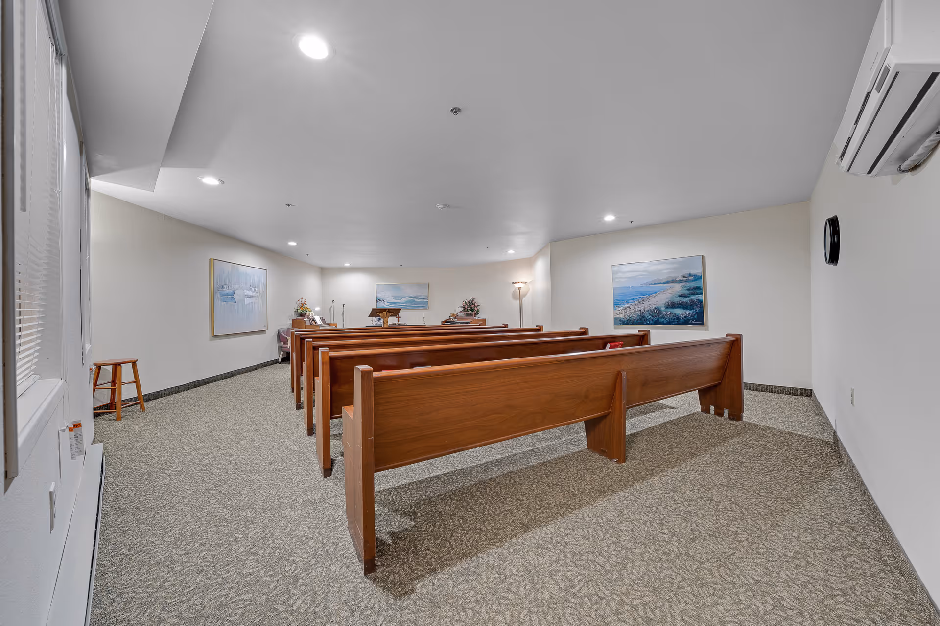 Carpeted chapel-style interior with rows of wooden pews facing a small podium and framed artwork on the walls.