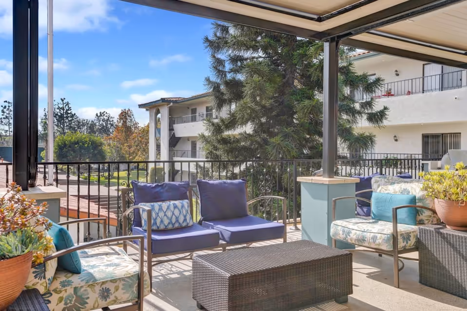 Covered outdoor patio with cushioned chairs, a wicker coffee table and potted plants overlooking a courtyard and neighboring building.