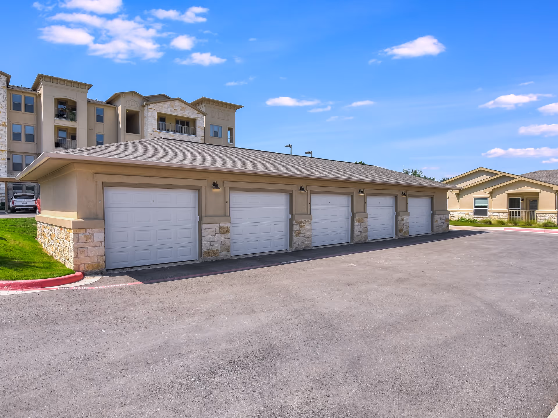 A row of five closed garage doors attached to a single-story building with beige walls and stone accents, situated in a senior living facility. In the background, there are multi-story residential buildings and a smaller building under a blue sky with scattered clouds.
