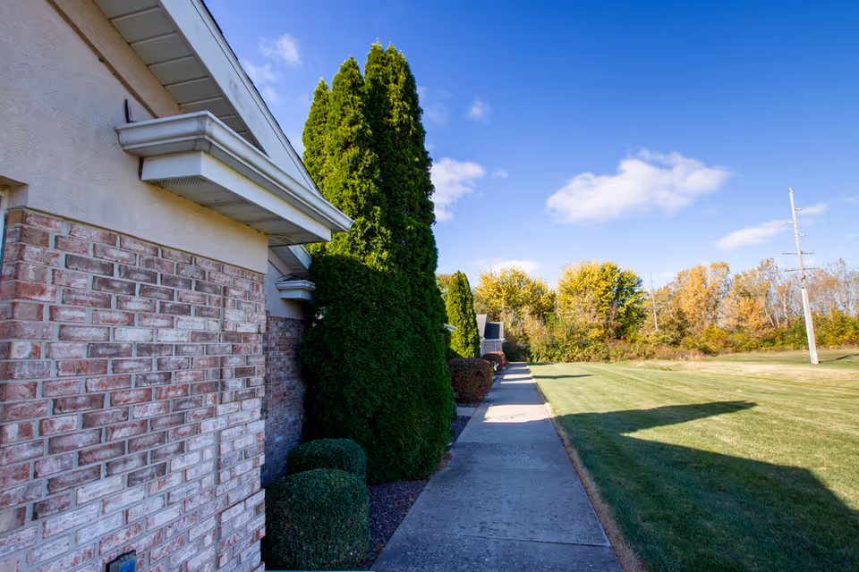 Side view of a building with brick and beige siding, a concrete walkway lined with tall green shrubs, and a grassy lawn extending to the right under a blue sky with scattered clouds.