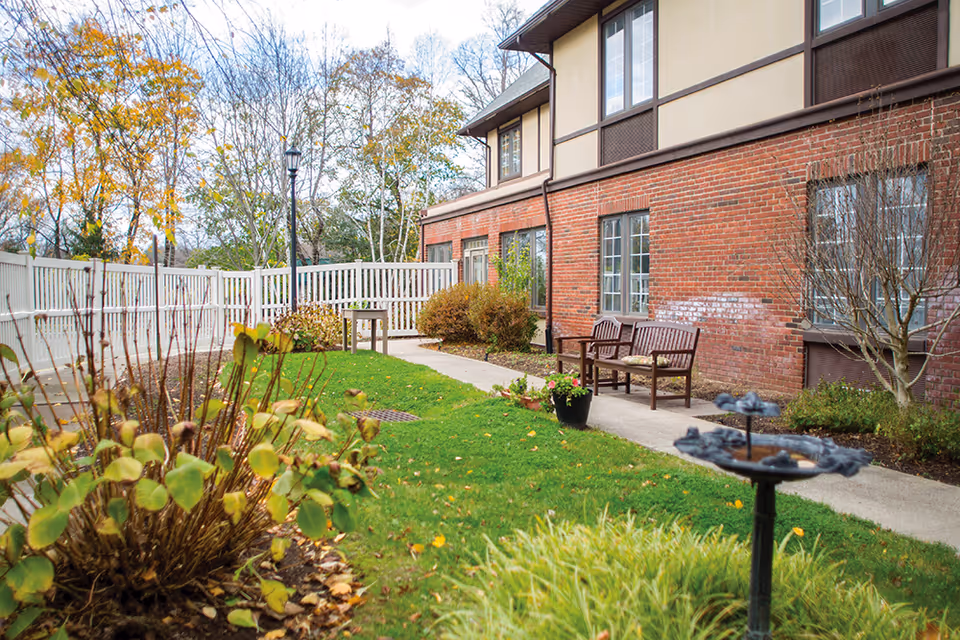 Outdoor garden area at Brookdale Florham Park featuring a green lawn, a paved walkway, wooden benches with cushions, a birdbath, and various plants and shrubs. The building has a brick exterior with multiple windows, and a white fence encloses the space. Trees with autumn foliage are visible in the background.