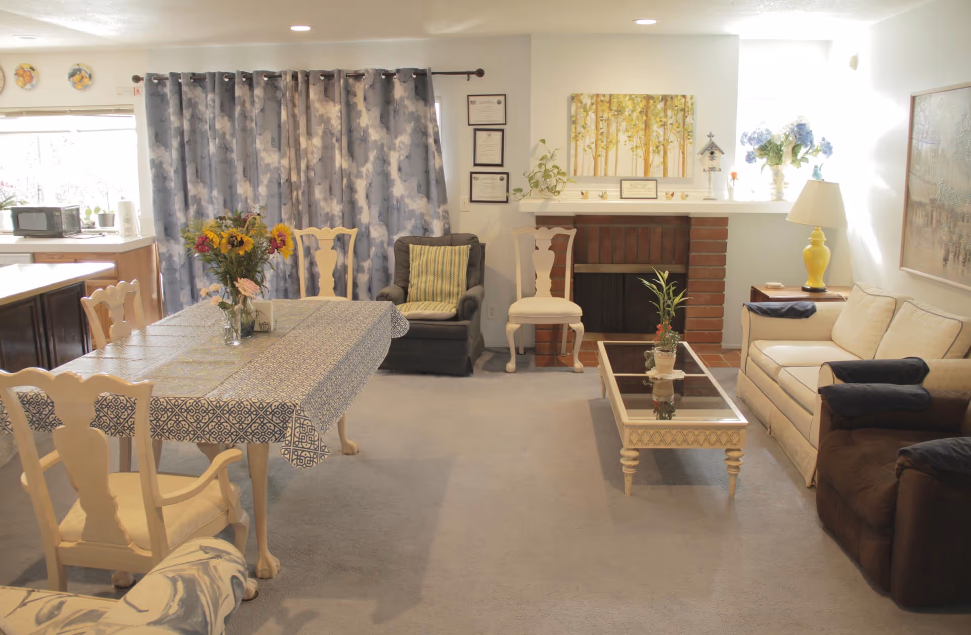 Bright combined living and dining room with a table and chairs in the foreground, sofas and armchairs around a coffee table, and a brick fireplace against the far wall.