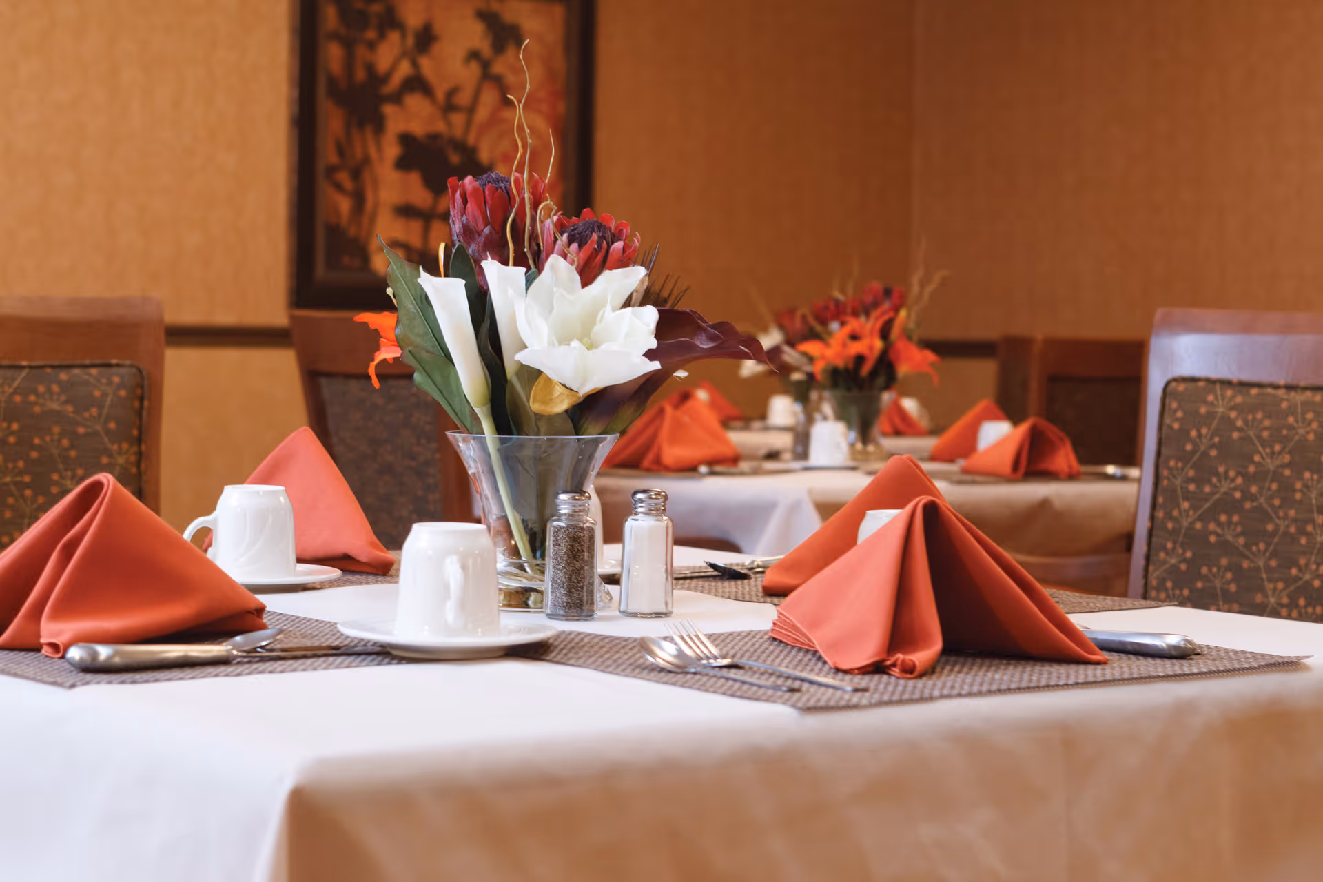A dining room table set with folded red napkins, white cups, a floral centerpiece, and salt and pepper shakers.