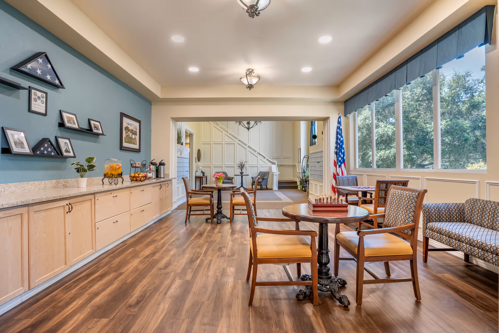A bright and spacious common area in a senior living facility with wooden flooring, multiple tables and chairs, a chess set on one table, large windows letting in natural light, an American flag, and a countertop with a beverage dispenser and fruit bowl. The walls are decorated with framed pictures and folded American flags in display cases.