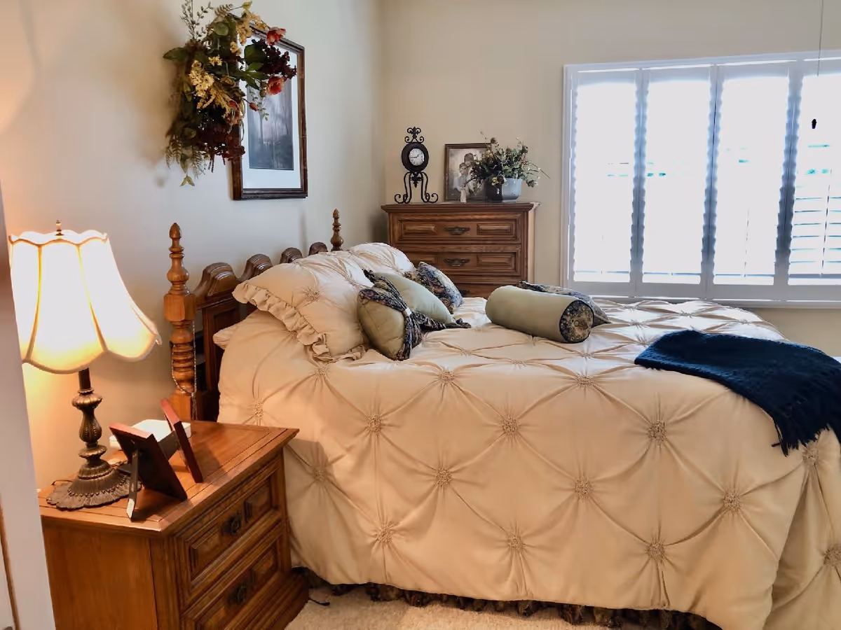 A cozy bedroom with a wooden bed frame and a cream-colored comforter adorned with decorative pillows. There is a wooden nightstand with a lamp and picture frames on the left side of the bed. A wooden dresser with decorative items and a framed photo is placed against the wall near a window with white plantation shutters. A floral wreath hangs on the wall above the bed.