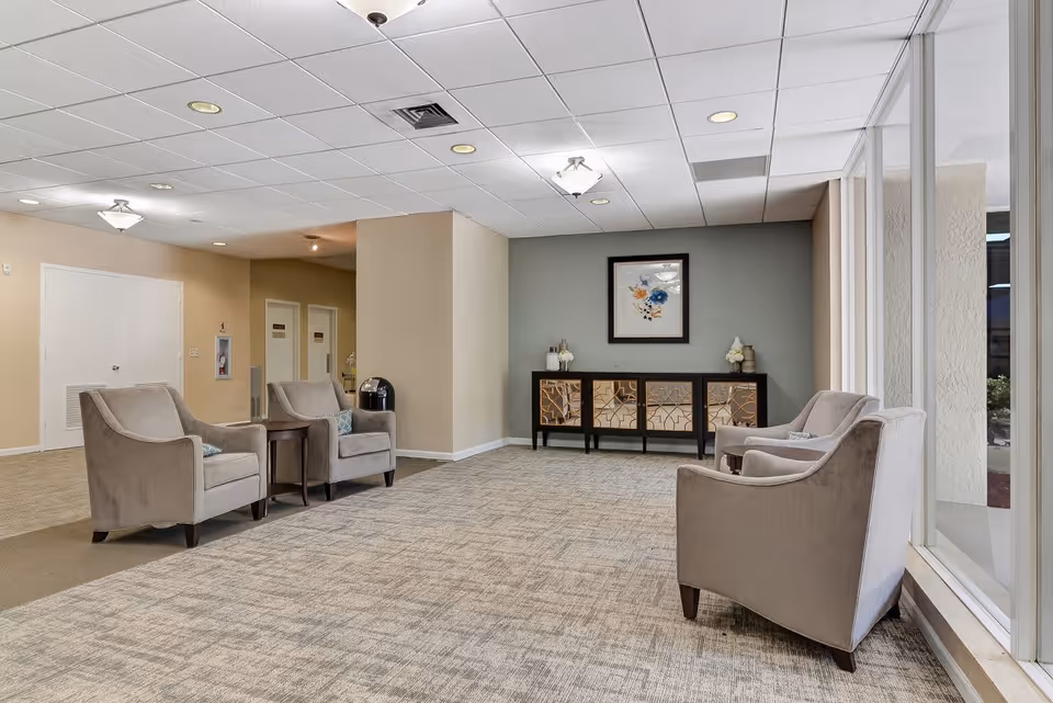 A spacious and well-lit lounge area with four beige armchairs arranged in pairs facing each other. There is a small wooden table between two of the chairs. Against the far wall is a decorative cabinet with mirrored panels and a framed floral artwork above it. Large windows on the right side let in natural light, and the ceiling has recessed lighting and a central light fixture.