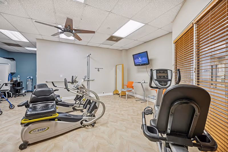 A fitness room with exercise equipment including recumbent bikes and elliptical machines. The room has light-colored wood flooring, a ceiling fan, a wall-mounted TV, and a window with wooden blinds. There are also two orange chairs and a small table in the corner.