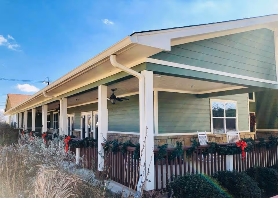 Exterior view of a single-story building with green siding and a covered porch decorated with garlands and red bows. The porch has white columns and rocking chairs, with plants and bushes in the foreground under a clear blue sky.
