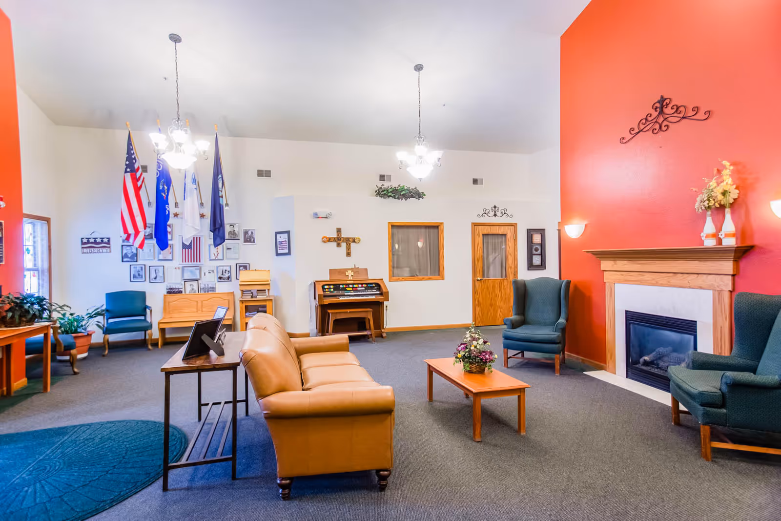 Communal living room in an assisted living facility with sofas, armchairs, a fireplace, flags, and wall decorations.