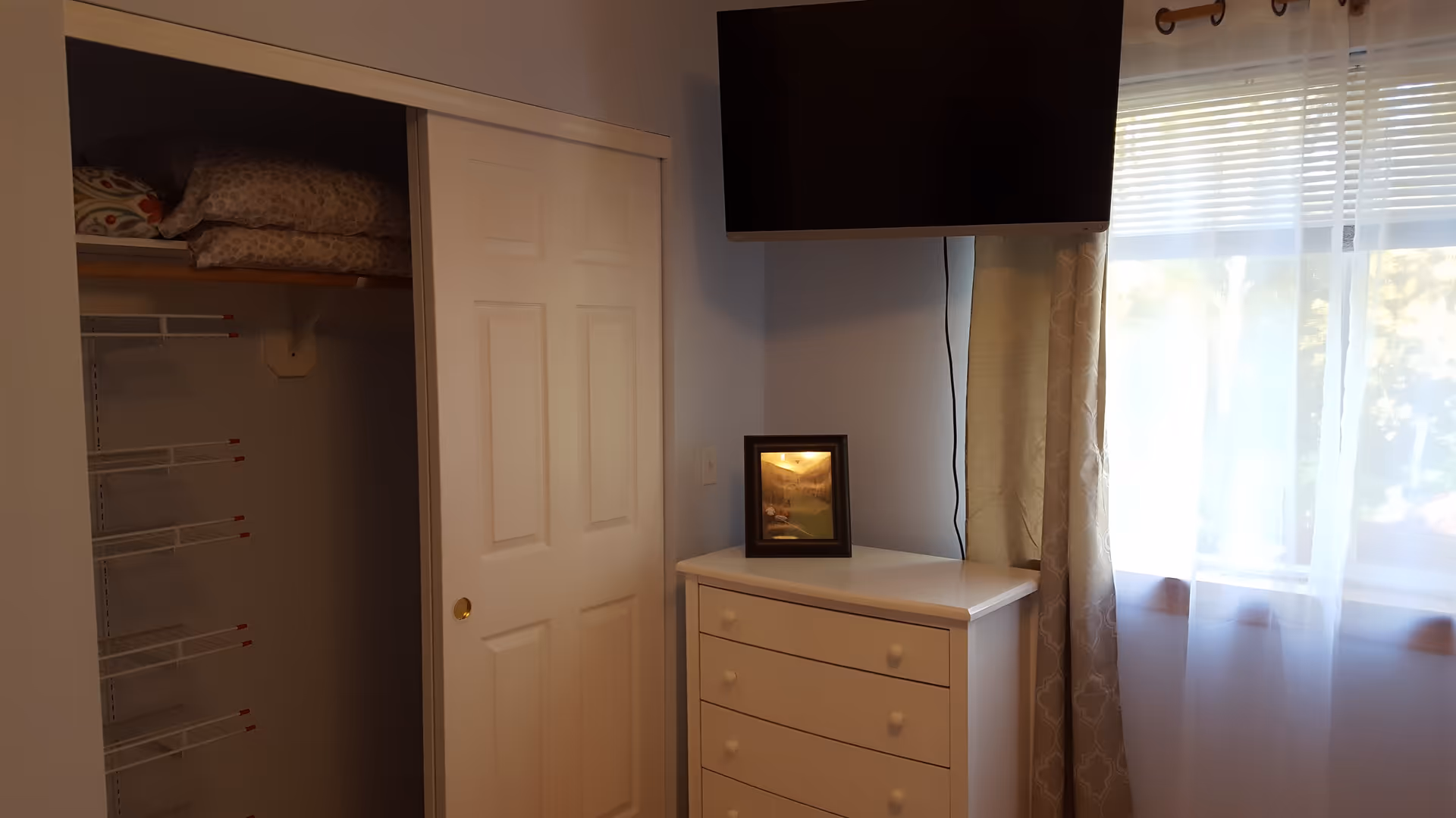 A corner of a bedroom featuring a white dresser with a framed picture on top, a wall-mounted flat-screen TV above it, a window with sheer and patterned curtains letting in natural light, and a partially open closet with shelves and folded bedding inside.