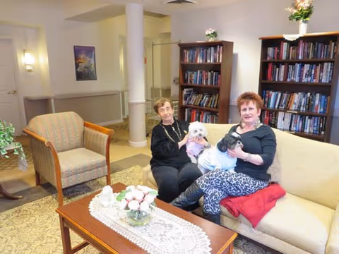Two elderly women sitting on a beige couch in a cozy room with bookshelves filled with books behind them. One woman is holding a small white dog, and the other woman is holding a small black dog. There is a wooden coffee table with a lace runner and a flower arrangement in front of them, and an armchair to the left. The room has soft lighting and a warm, welcoming atmosphere.