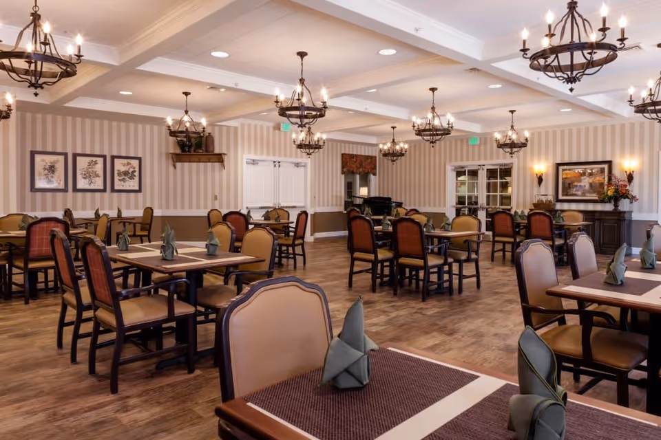 Well-lit dining room with multiple tables and chairs, chandeliers, and neatly folded napkins on wood floors.